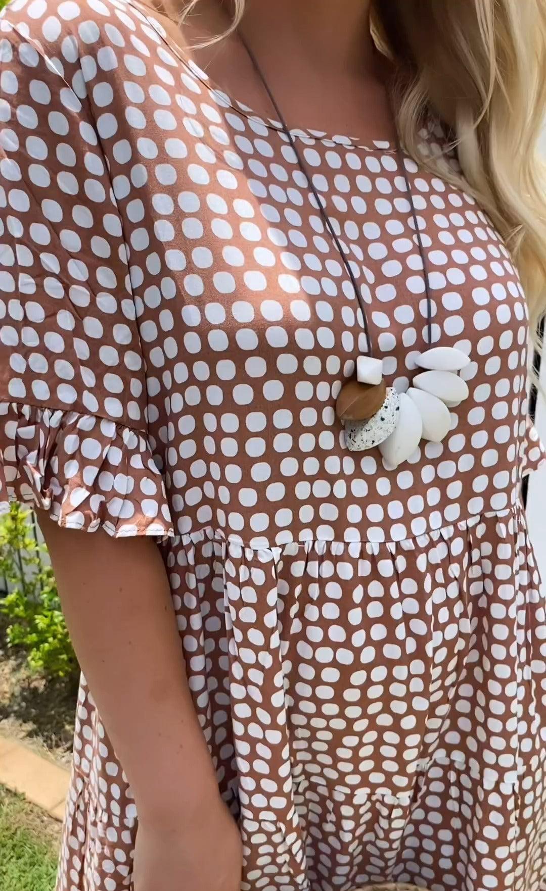 Red - A woman wearing a brown and white dot print tiered midi dress with ruffle-cuffed elbow sleeves, standing outdoors near a white picket fence.