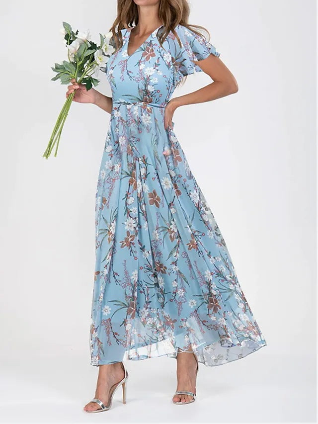 Blue - A woman wearing a dusty blue floral print chiffon maxi dress with flutter sleeves, a V-neckline, and a flowing skirt, paired with silver heeled sandals.