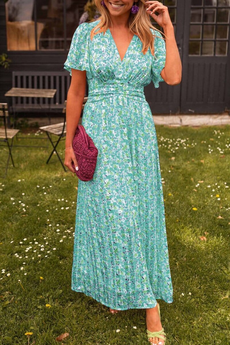 Green - A woman wearing a vibrant orange and pink floral print maxi dress with flutter sleeves, a plunging V-neckline, and a ruched waistband, standing on a green lawn.
