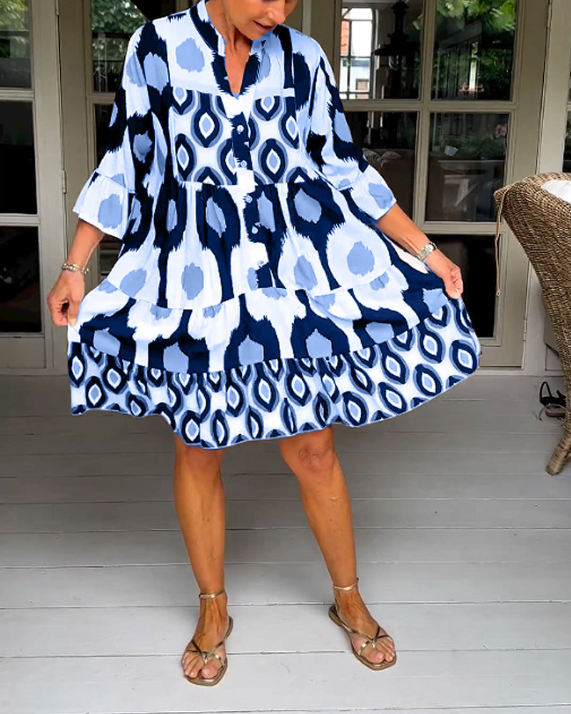 Blue - A woman wearing a knee-length, tiered tunic dress with a black, tan, and white geometric ikat print, featuring elbow-length sleeves with ruffled cuffs and a split V-neckline.