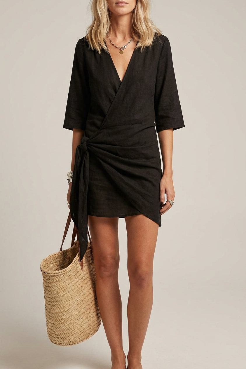 Black - A woman wearing a light oatmeal-colored textured linen wrap mini dress with elbow-length sleeves, a deep V-neck, and a side-tie knot, paired with a woven straw bag.