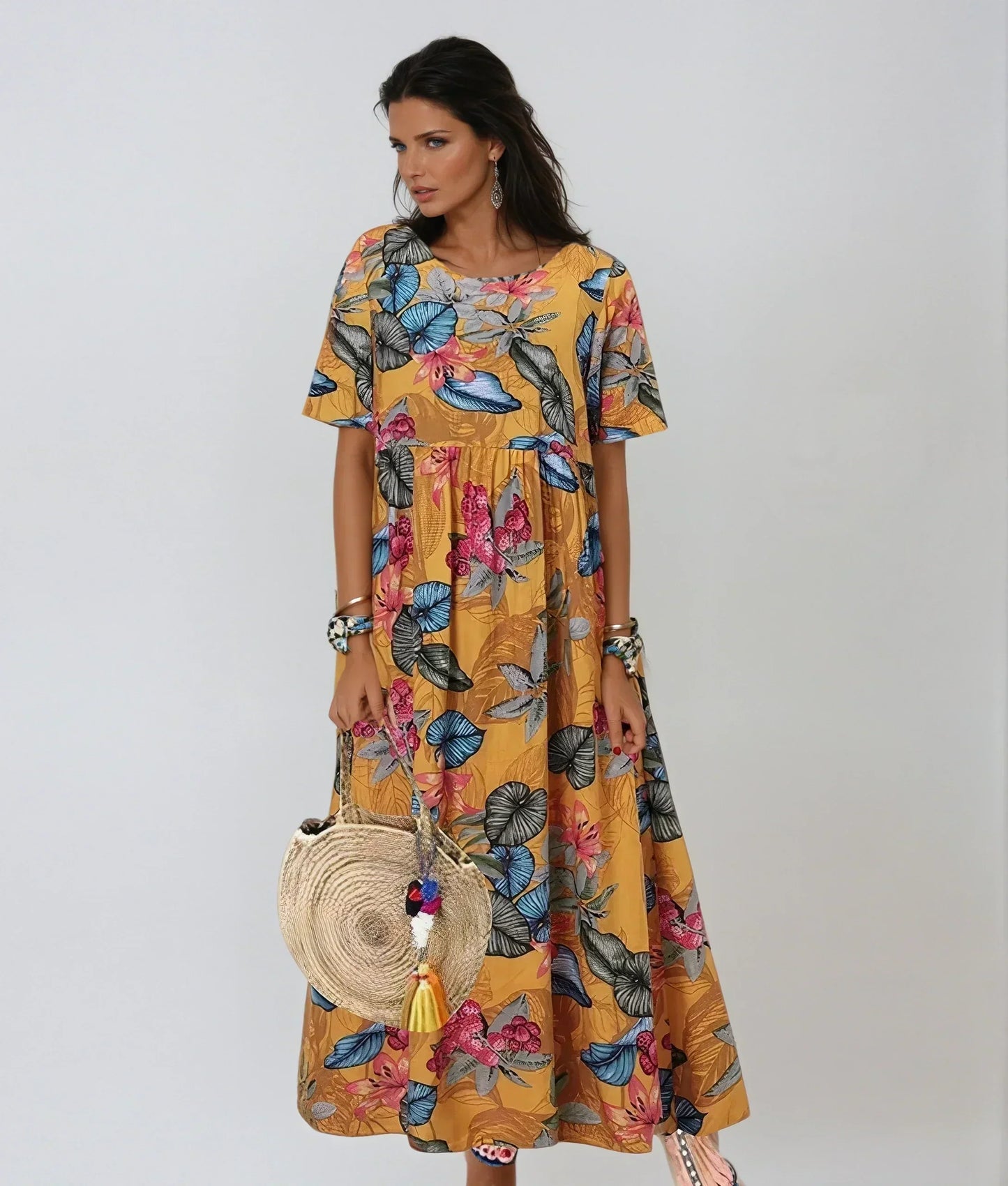 Yellow - A woman wearing a navy blue maxi dress with a vibrant pink, orange, and green tropical floral print, featuring short sleeves, a crewneck, and an empire waist, holding a round woven straw bag.