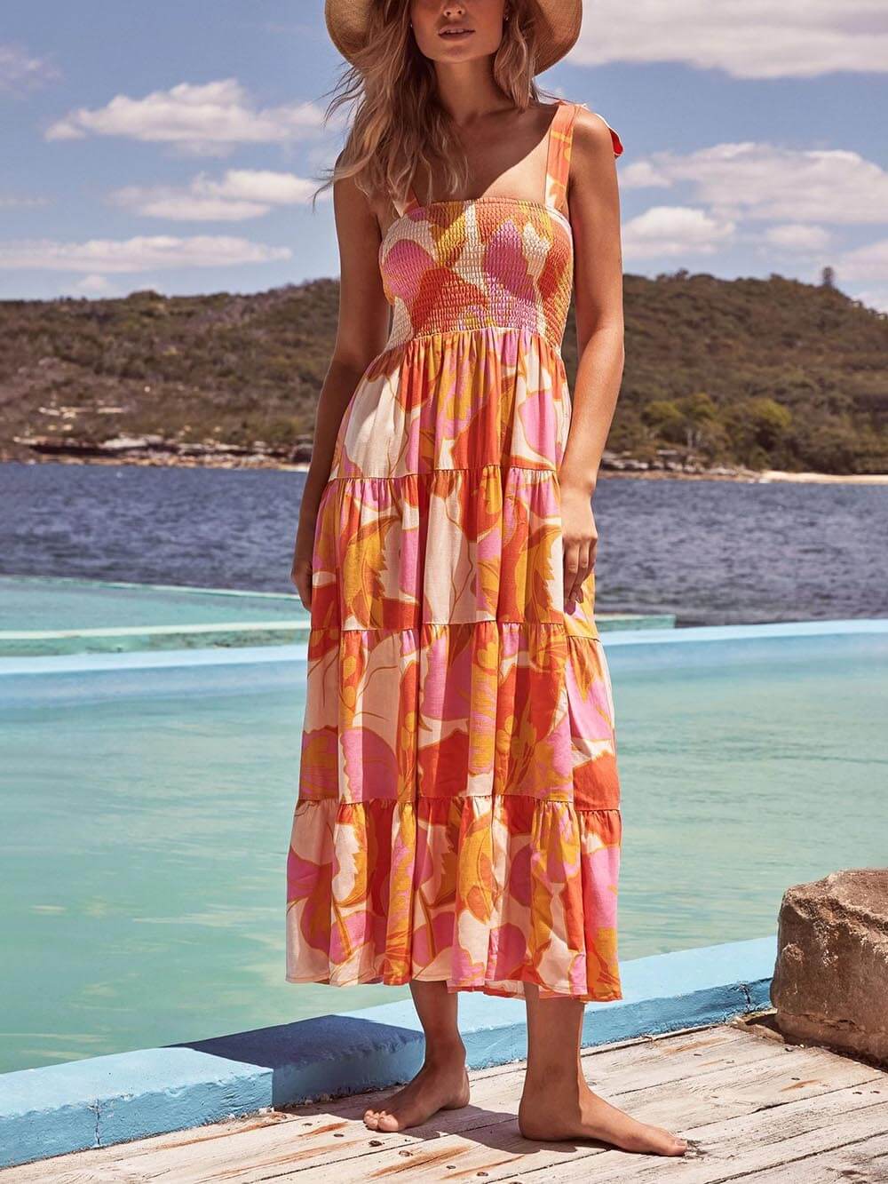 A woman wearing a sleeveless, tiered maxi dress with a pink and orange abstract floral print, featuring a smocked bodice and tie straps, standing on a wooden deck by the water.