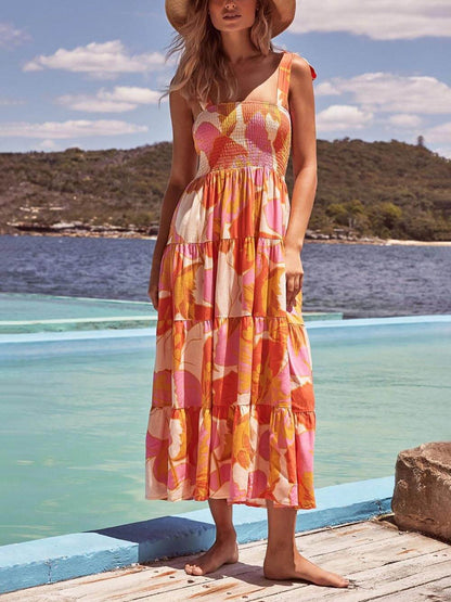 A woman wearing a sleeveless, tiered maxi dress with a pink and orange abstract floral print, featuring a smocked bodice and tie straps, standing on a wooden deck by the water.