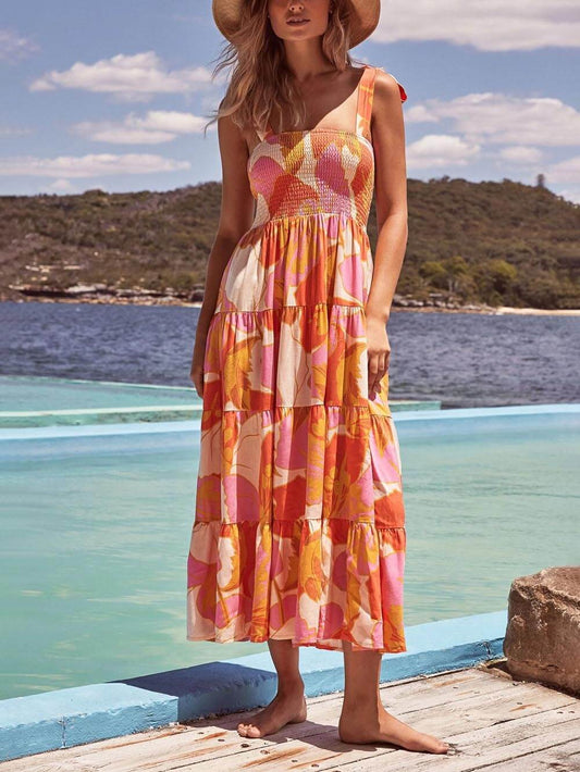 A woman wearing a sleeveless, tiered maxi dress with a pink and orange abstract floral print, featuring a smocked bodice and tie straps, standing on a wooden deck by the water.