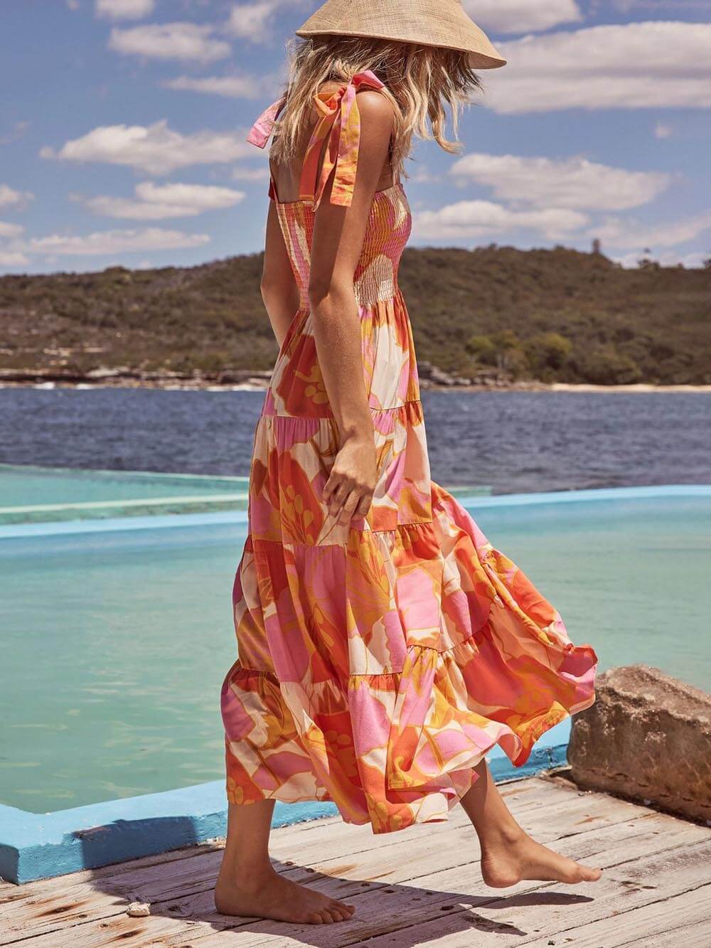 A woman wearing a sleeveless, tiered maxi dress with a pink and orange abstract floral print, featuring a smocked bodice and tie straps, standing on a wooden deck by the water.