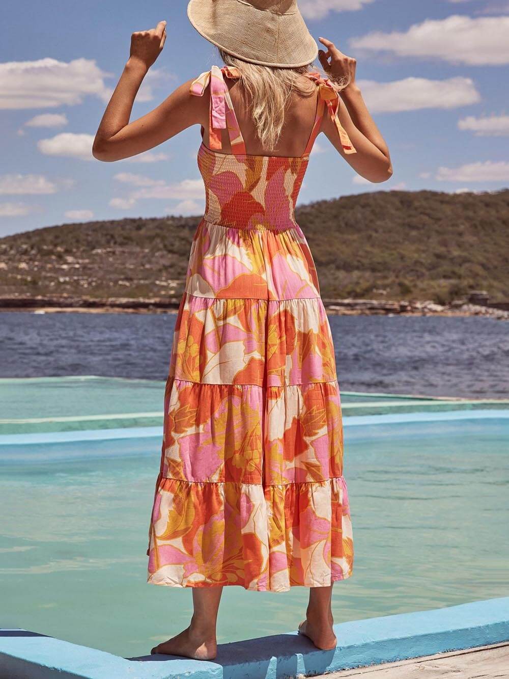 A woman wearing a sleeveless, tiered maxi dress with a pink and orange abstract floral print, featuring a smocked bodice and tie straps, standing on a wooden deck by the water.