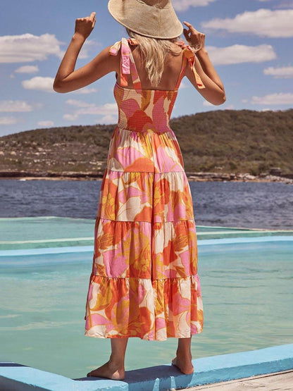 A woman wearing a sleeveless, tiered maxi dress with a pink and orange abstract floral print, featuring a smocked bodice and tie straps, standing on a wooden deck by the water.