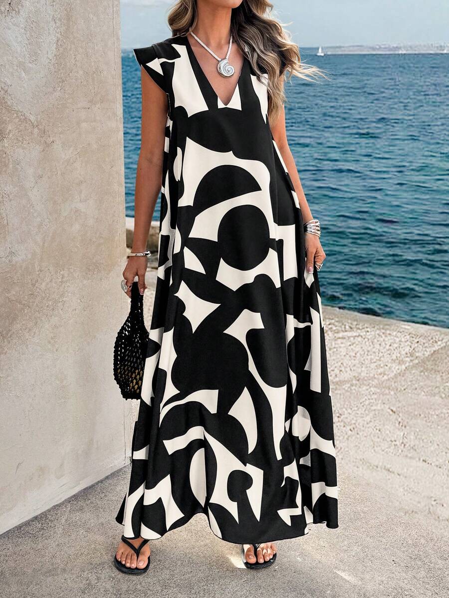 Black - A woman wearing a sleeveless, relaxed-fit maxi dress with a bold black and white abstract graphic print, featuring a V-neckline, standing near a stone wall with the blue ocean in the background.