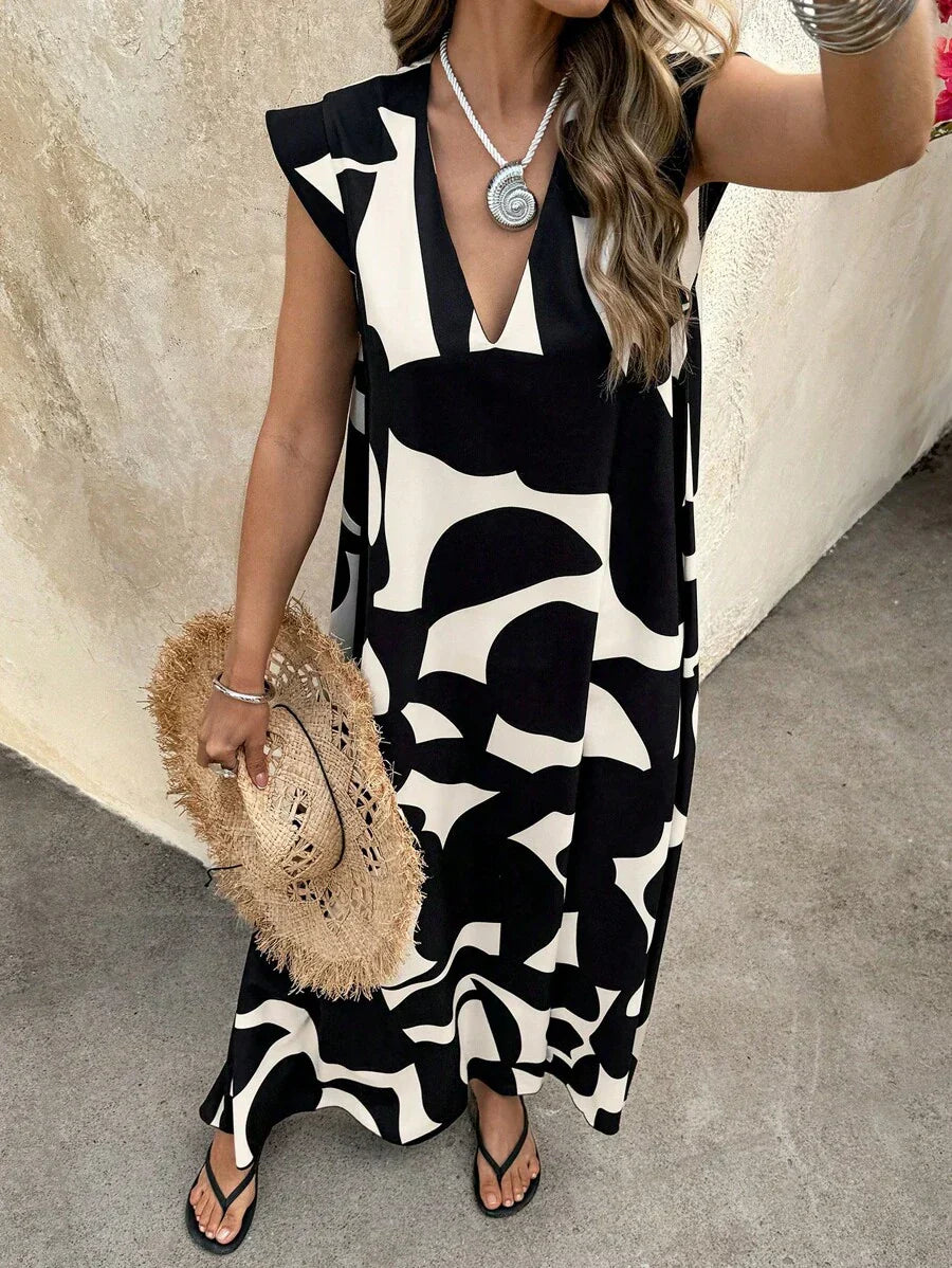 Black - A woman wearing a sleeveless, relaxed-fit maxi dress with a bold black and white abstract graphic print, featuring a V-neckline, standing near a stone wall with the blue ocean in the background.
