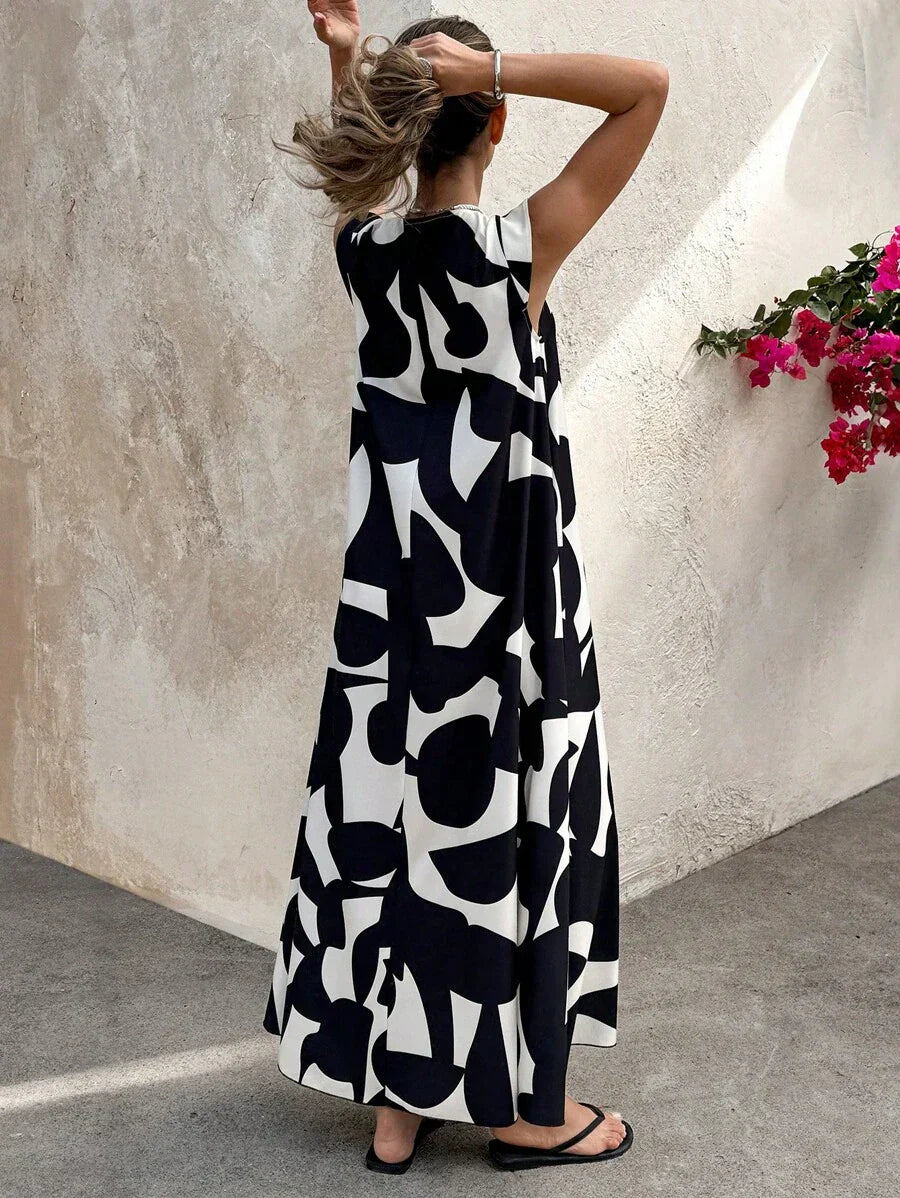 Black - A woman wearing a sleeveless, relaxed-fit maxi dress with a bold black and white abstract graphic print, featuring a V-neckline, standing near a stone wall with the blue ocean in the background.