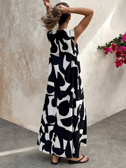 Black - A woman wearing a sleeveless, relaxed-fit maxi dress with a bold black and white abstract graphic print, featuring a V-neckline, standing near a stone wall with the blue ocean in the background.