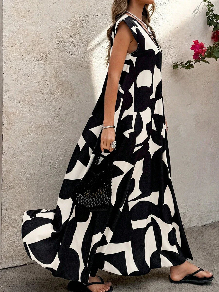 Black - A woman wearing a sleeveless, relaxed-fit maxi dress with a bold black and white abstract graphic print, featuring a V-neckline, standing near a stone wall with the blue ocean in the background.