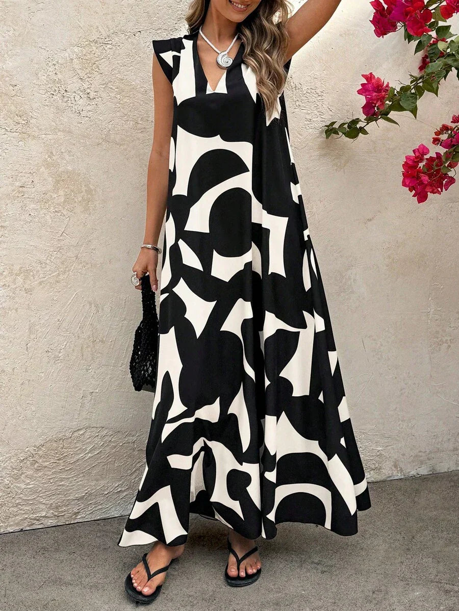 Black - A woman wearing a sleeveless, relaxed-fit maxi dress with a bold black and white abstract graphic print, featuring a V-neckline, standing near a stone wall with the blue ocean in the background.