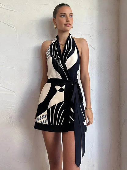 Woman wearing a black and white abstract geometric crepe mini dress featuring a halter neckline, wrap bodice, and long waist tie sash.