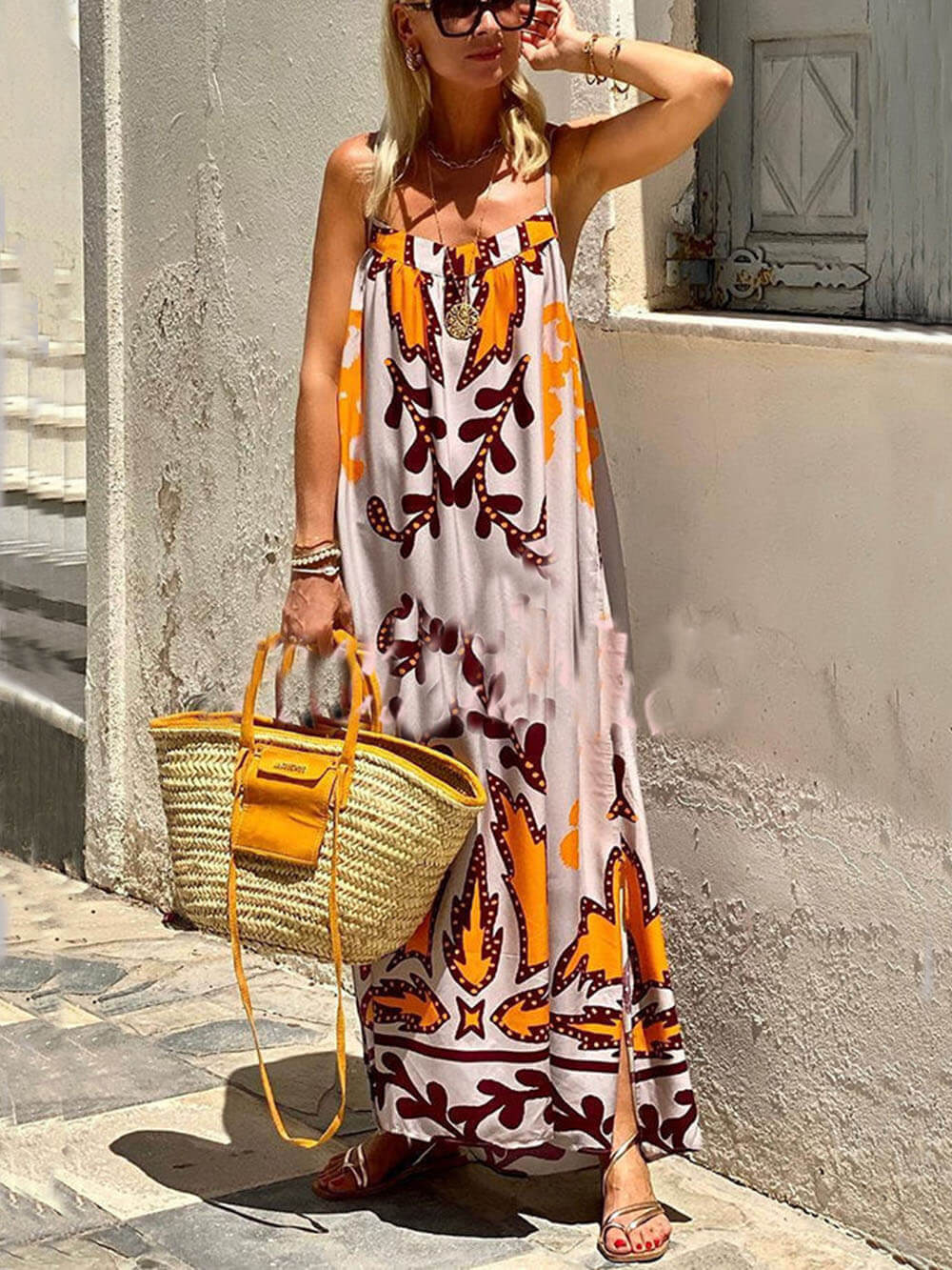 A woman wearing a floor-length sleeveless maxi dress with a bold orange and brown abstract leaf print on a light grey background, holding a woven straw tote bag.