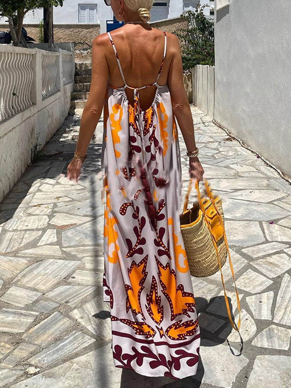 A woman wearing a floor-length sleeveless maxi dress with a bold orange and brown abstract leaf print on a light grey background, holding a woven straw tote bag.