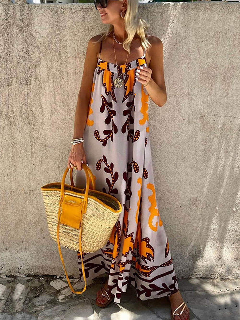 Multicolor - A woman wearing a floor-length sleeveless maxi dress with a bold orange and brown abstract leaf print on a light grey background, holding a woven straw tote bag.