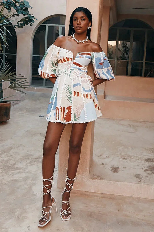 Woman wearing an abstract print off-the-shoulder mini dress with puff sleeves, a notched neckline, and crochet trim detailing, standing in a sunlit courtyard.