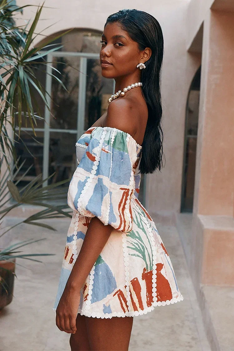 sand - Woman wearing an abstract print off-the-shoulder mini dress with puff sleeves, a notched neckline, and crochet trim detailing, standing in a sunlit courtyard.