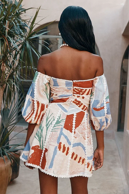 sand - Woman wearing an abstract print off-the-shoulder mini dress with puff sleeves, a notched neckline, and crochet trim detailing, standing in a sunlit courtyard.