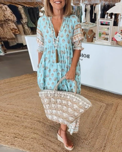 Blue - A woman wearing a light blue block-print maxi dress with a split V-neckline, tassel ties, and tiered skirt, holding a woven straw tote bag.