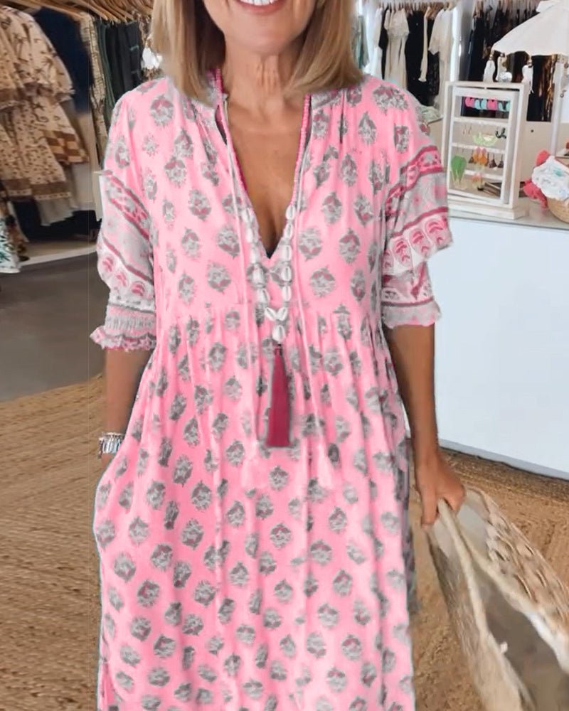 Pink - A woman wearing a light blue block-print maxi dress with a split V-neckline, tassel ties, and tiered skirt, holding a woven straw tote bag.