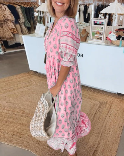 Pink - A woman wearing a light blue block-print maxi dress with a split V-neckline, tassel ties, and tiered skirt, holding a woven straw tote bag.