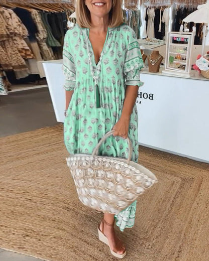 Green - A woman wearing a light blue block-print maxi dress with a split V-neckline, tassel ties, and tiered skirt, holding a woven straw tote bag.