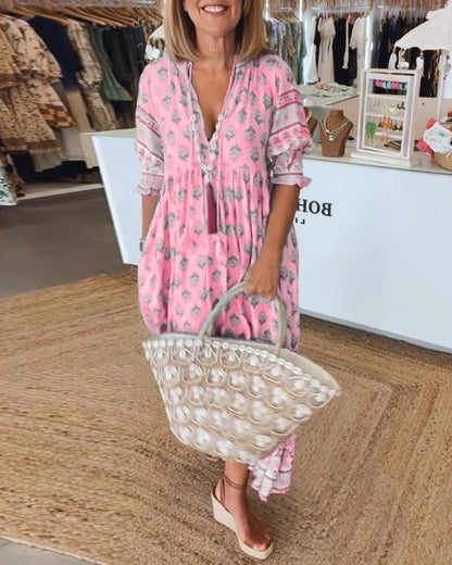 Pink - A woman wearing a light blue block-print maxi dress with a split V-neckline, tassel ties, and tiered skirt, holding a woven straw tote bag.