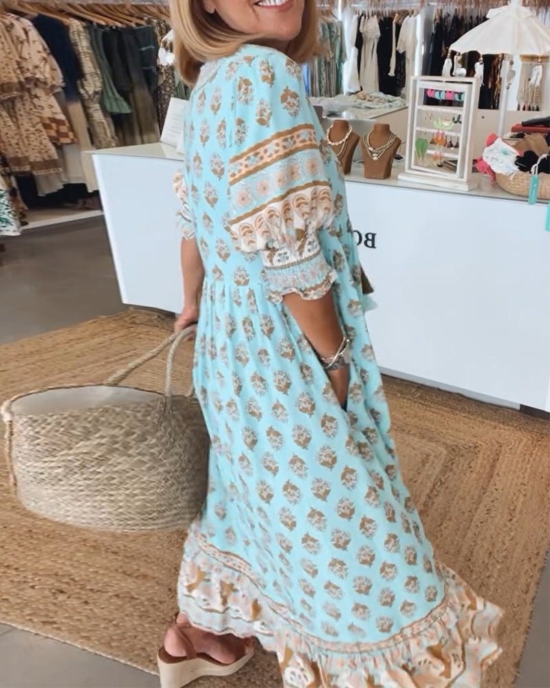 Pink - A woman wearing a light blue block-print maxi dress with a split V-neckline, tassel ties, and tiered skirt, holding a woven straw tote bag.