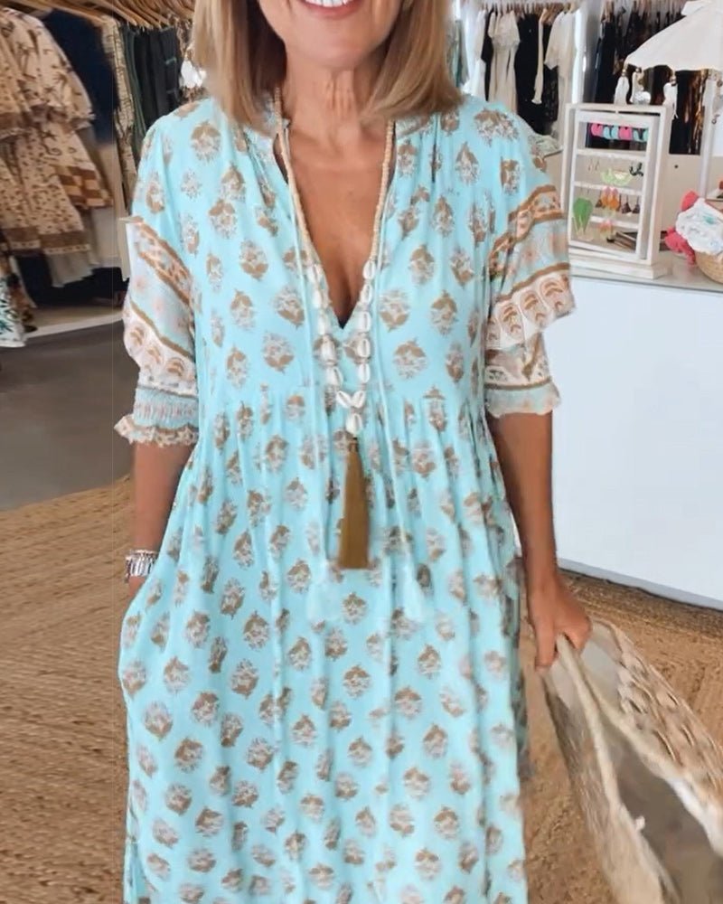 Pink - A woman wearing a light blue block-print maxi dress with a split V-neckline, tassel ties, and tiered skirt, holding a woven straw tote bag.