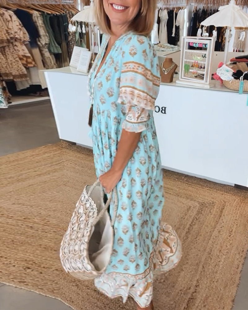 Pink - A woman wearing a light blue block-print maxi dress with a split V-neckline, tassel ties, and tiered skirt, holding a woven straw tote bag.