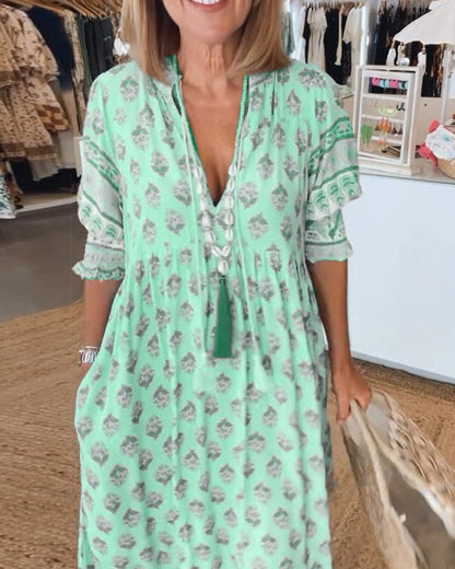 Pink - A woman wearing a light blue block-print maxi dress with a split V-neckline, tassel ties, and tiered skirt, holding a woven straw tote bag.