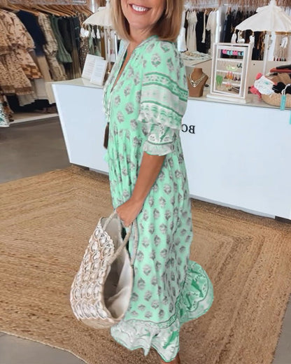 Pink - A woman wearing a light blue block-print maxi dress with a split V-neckline, tassel ties, and tiered skirt, holding a woven straw tote bag.