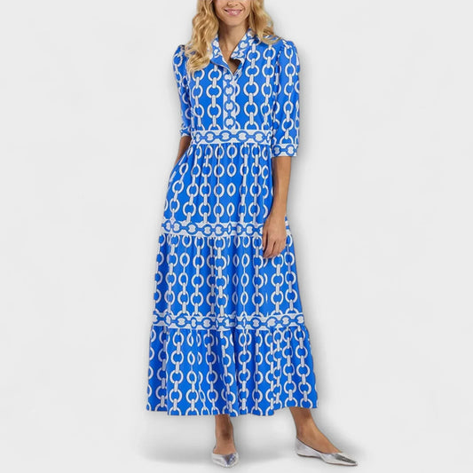 A woman wearing a royal blue and white chain-link print maxi dress featuring a pointed collar, elbow-length sleeves, a defined waist, and a tiered skirt, paired with silver pointed-toe flats.