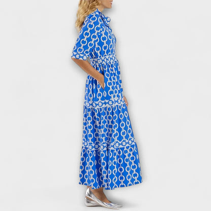 A woman wearing a royal blue and white chain-link print maxi dress featuring a pointed collar, elbow-length sleeves, a defined waist, and a tiered skirt, paired with silver pointed-toe flats.