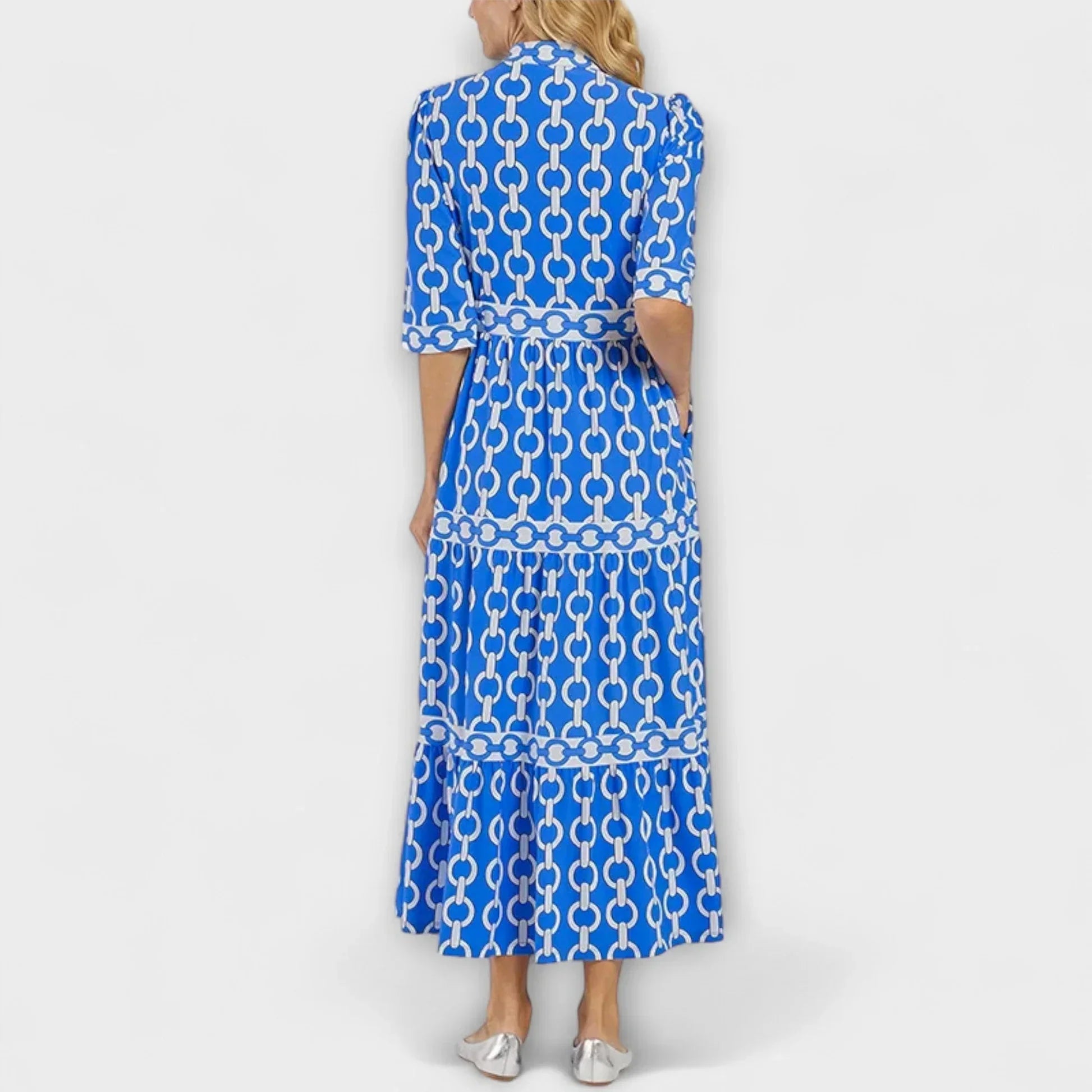 A woman wearing a royal blue and white chain-link print maxi dress featuring a pointed collar, elbow-length sleeves, a defined waist, and a tiered skirt, paired with silver pointed-toe flats.