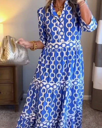 Blue - A woman wearing a royal blue and white chain-link print maxi dress featuring a pointed collar, elbow-length sleeves, a defined waist, and a tiered skirt, paired with silver pointed-toe flats.