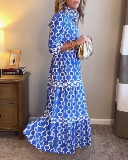 Blue - A woman wearing a royal blue and white chain-link print maxi dress featuring a pointed collar, elbow-length sleeves, a defined waist, and a tiered skirt, paired with silver pointed-toe flats.