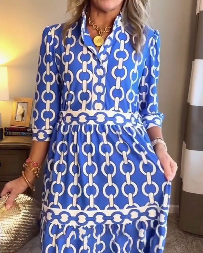 Blue - A woman wearing a royal blue and white chain-link print maxi dress featuring a pointed collar, elbow-length sleeves, a defined waist, and a tiered skirt, paired with silver pointed-toe flats.