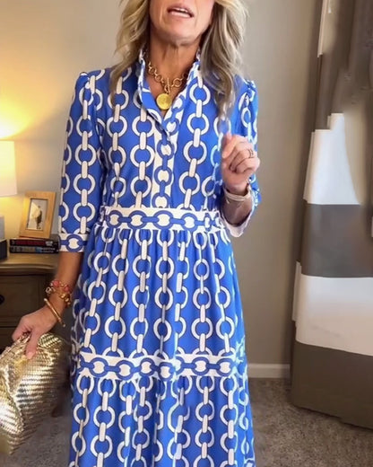 Blue - A woman wearing a royal blue and white chain-link print maxi dress featuring a pointed collar, elbow-length sleeves, a defined waist, and a tiered skirt, paired with silver pointed-toe flats.