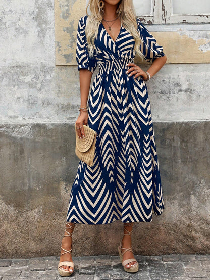 Blue - A woman wearing a navy and cream chevron print maxi dress with a smocked waist, V-neckline, puff sleeves, and a high side slit, paired with lace-up sandals.