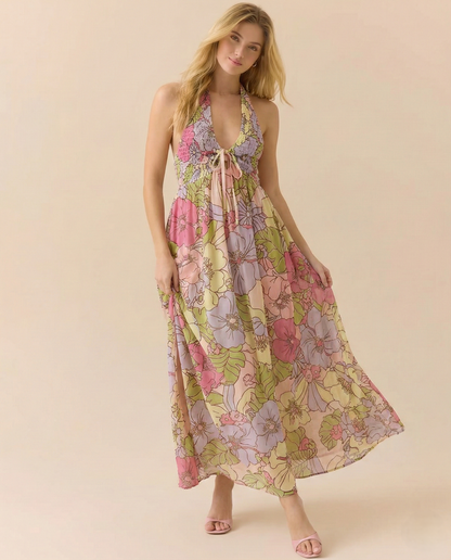 Garden - A woman wearing a pastel floral print chiffon maxi dress with a smocked halter bodice, tie-neck detail, and a flowing skirt with a side slit.