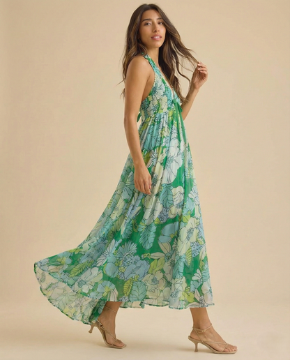 Ivy - A woman wearing a pastel floral print chiffon maxi dress with a smocked halter bodice, tie-neck detail, and a flowing skirt with a side slit.