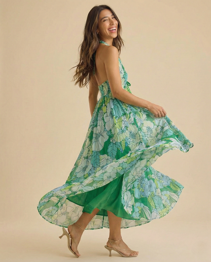 Ivy - A woman wearing a pastel floral print chiffon maxi dress with a smocked halter bodice, tie-neck detail, and a flowing skirt with a side slit.