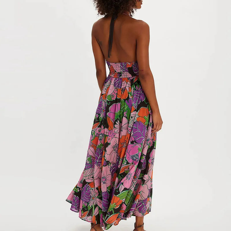 Ivy - A woman wearing a pastel floral print chiffon maxi dress with a smocked halter bodice, tie-neck detail, and a flowing skirt with a side slit.