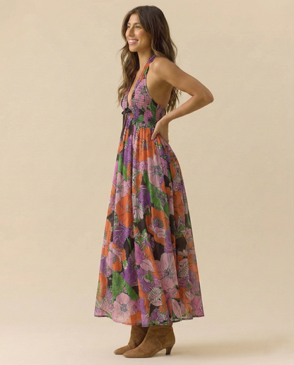 Tropical - A woman wearing a pastel floral print chiffon maxi dress with a smocked halter bodice, tie-neck detail, and a flowing skirt with a side slit.