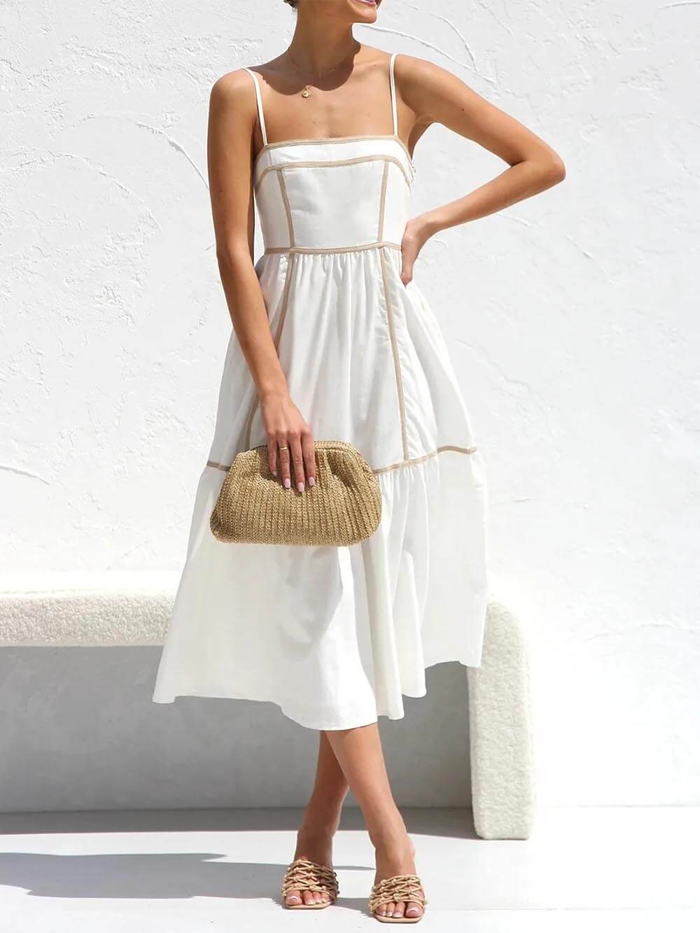 A woman wearing a white cotton poplin midi dress with tan contrast piping along the paneled bodice and tiered skirt, holding a woven straw clutch.