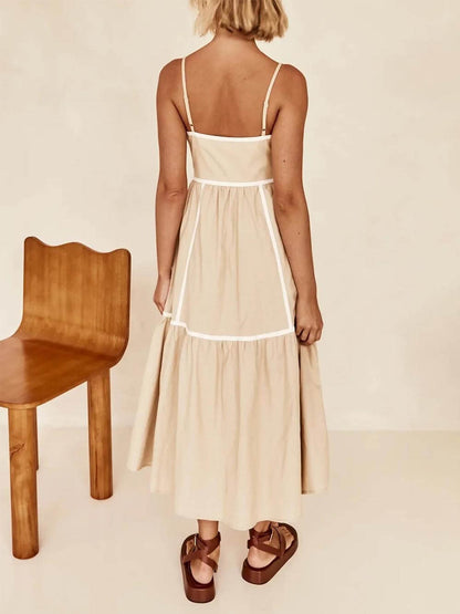 A woman wearing a white cotton poplin midi dress with tan contrast piping along the paneled bodice and tiered skirt, holding a woven straw clutch.
